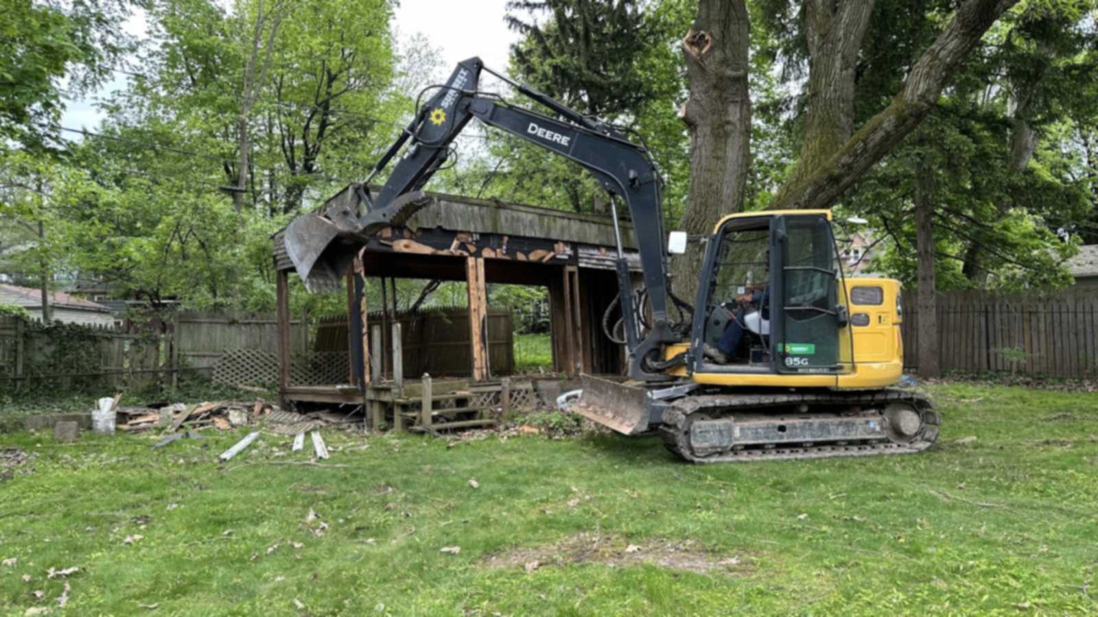 A tracked John Deere excavator with its bucket raised against an old wooden deck and roof structure in a tree-lined backyard.