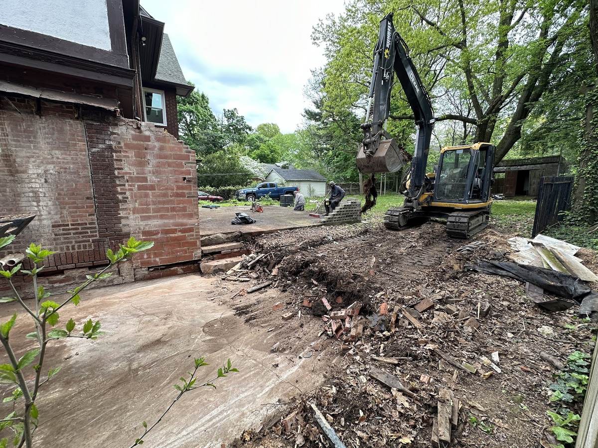 A tracked John Deere excavator on a cleared lot beside a brick house wall, with two workers breaking up a slab and brick, wood and soil in the foreground.