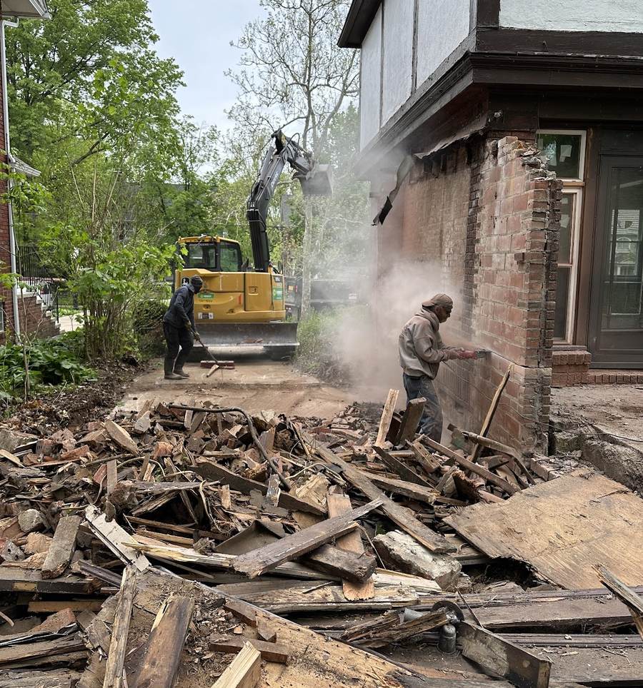 A worker cutting into a brick wall with a saw in a cloud of dust beside a pile of broken lumber, with a second worker and a yellow excavator behind him.