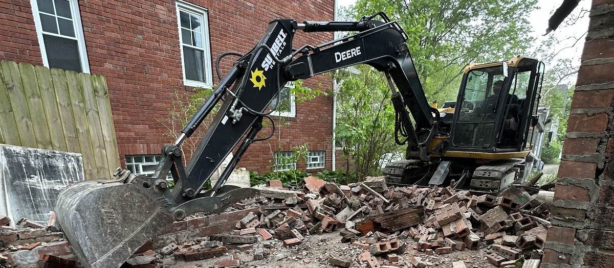 A tracked excavator with its bucket lowered among piles of broken brick beside a red brick house.
